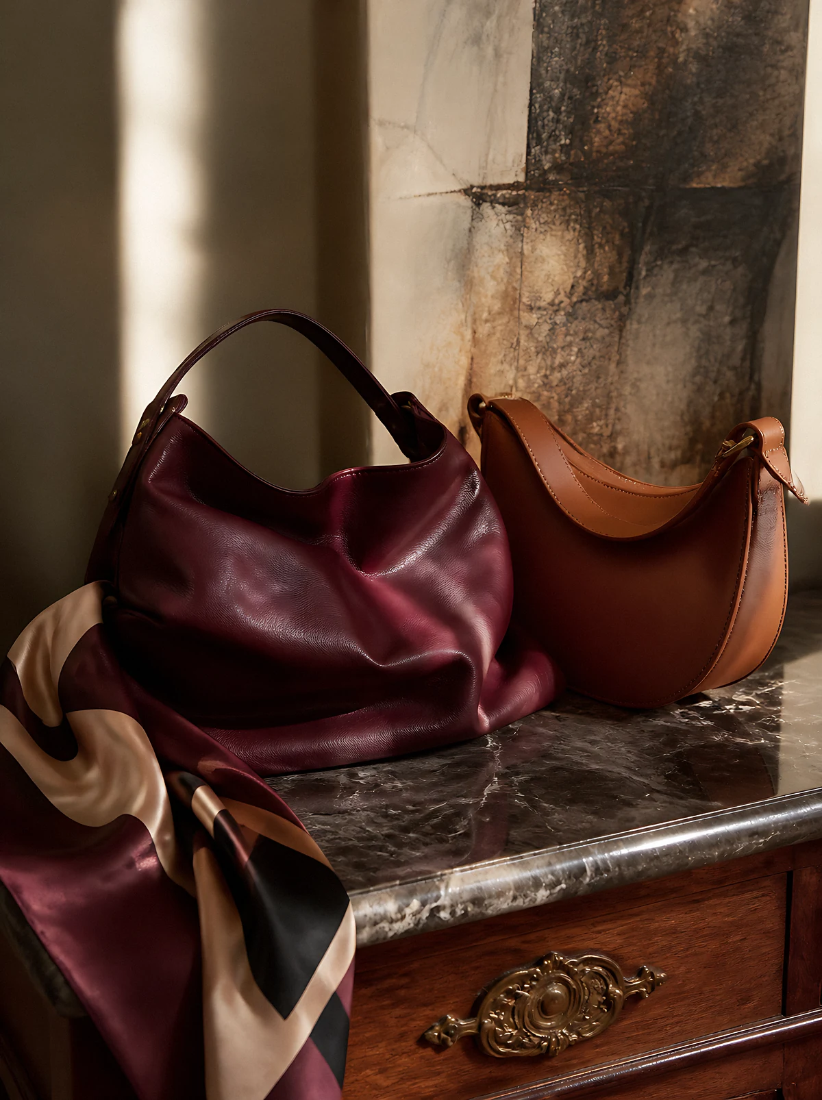 A slouchy oxblood leather hobo bag and a cognac half-moon bag on a marble console beside a folded silk scarf