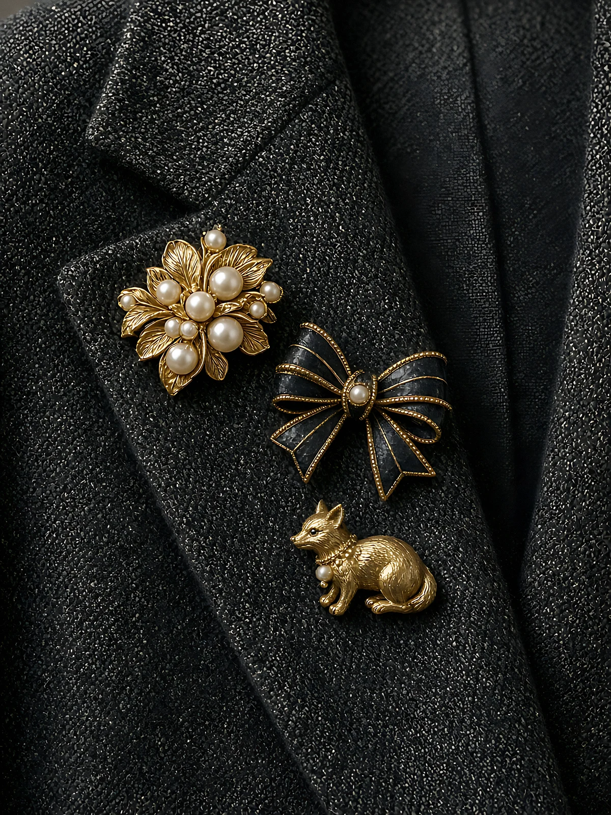 Three vintage-style brooches — a pearl flower, an enamel bow and a small gold animal — pinned to a tweed lapel