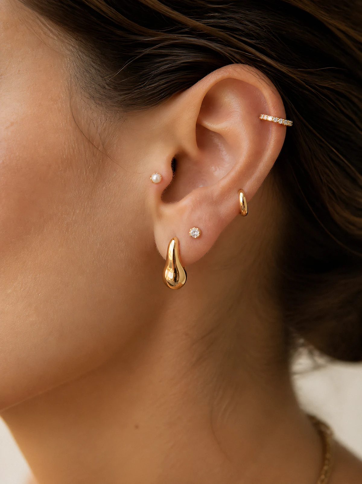 A close-up of an ear with a curated stack of gold jewellery: a sculptural drop, a tiny diamond stud, a huggie hoop, a helix cuff and a tragus stud