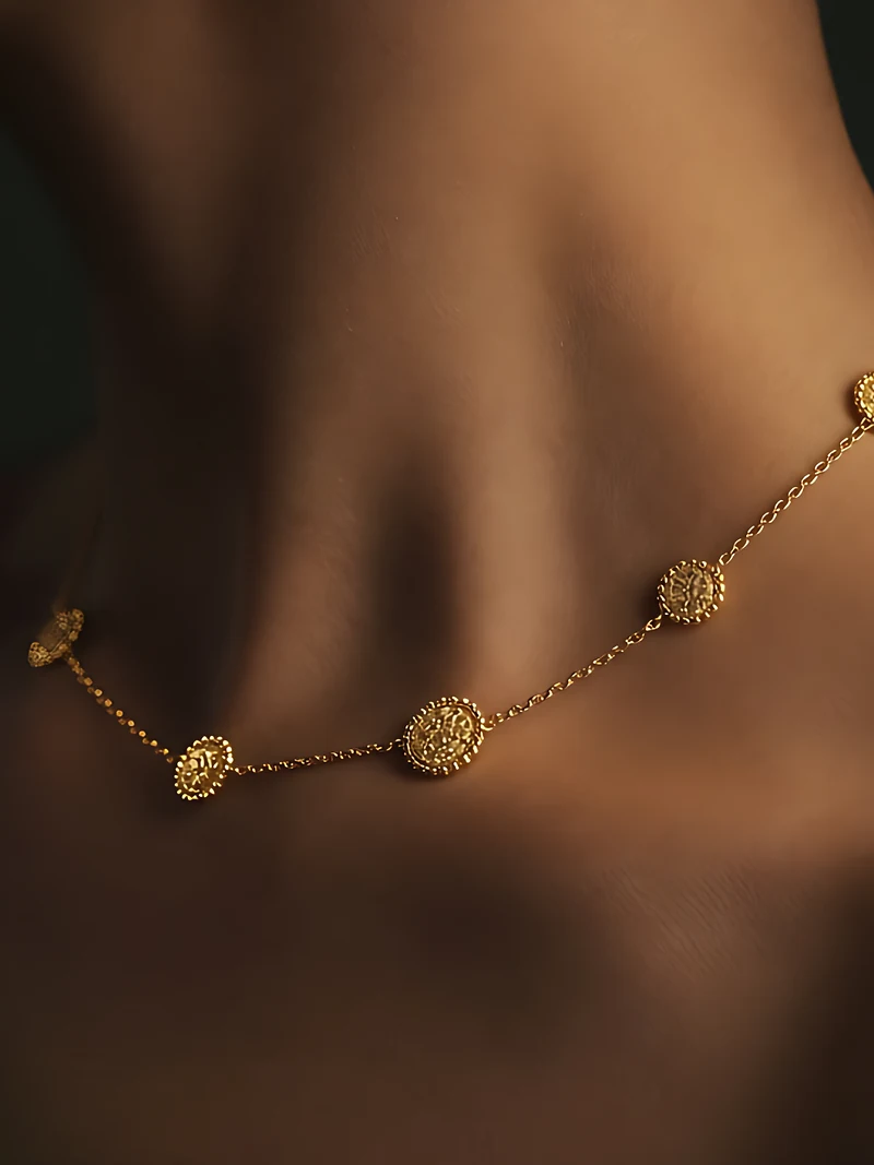 A fine gold chain of small round beaded medallions lying across a bare collarbone in shadow