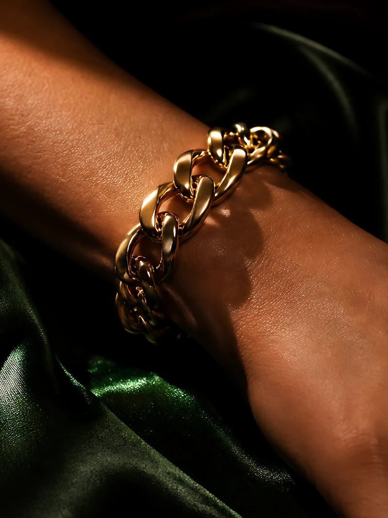 A bare wrist on dark green satin wearing a bold chain bracelet of rounded gold links, caught in a single beam of warm light