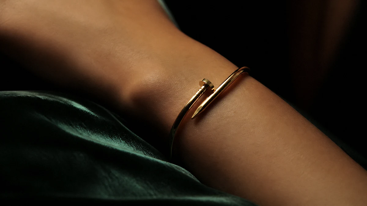 A bare wrist in low warm light wearing a gold bracelet shaped like a bent nail, resting on dark green satin