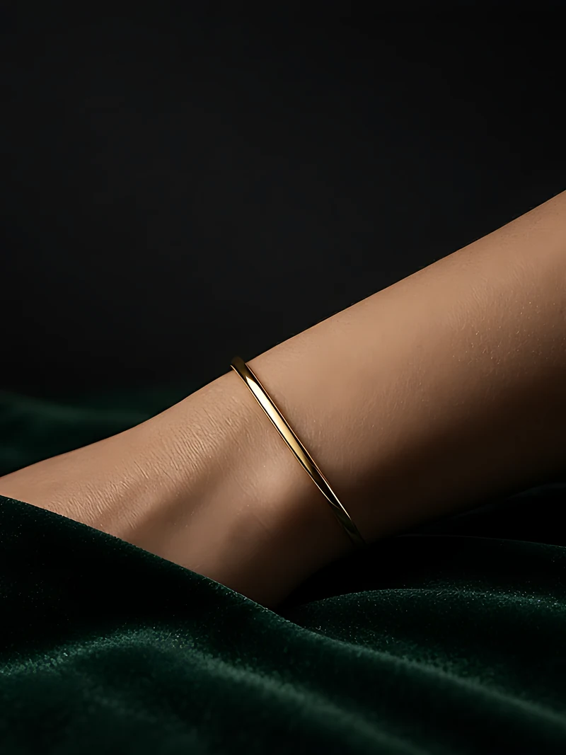 A wrist resting on dark green velvet, circled by a slim oval gold bangle in low, warm light
