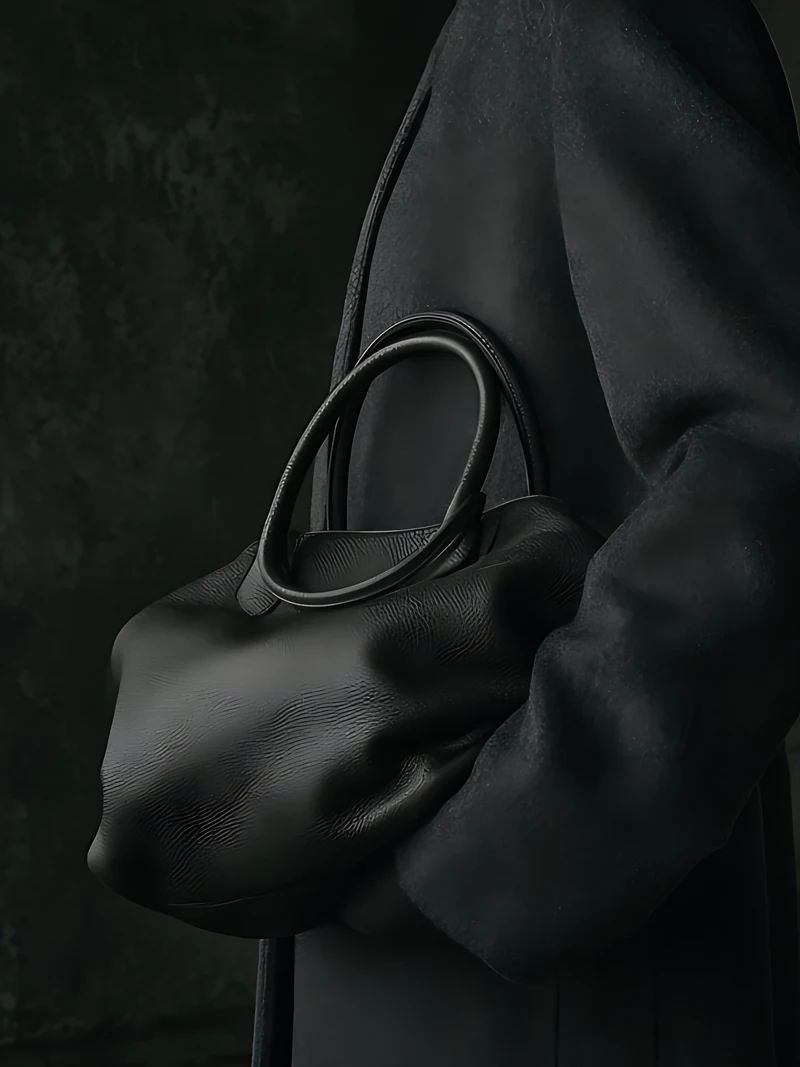 A figure in a long charcoal coat holding a soft black top-handle bag in a dim stone room