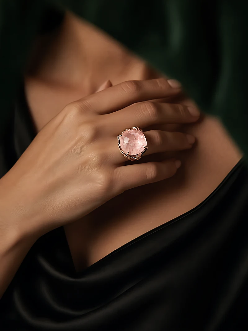 A hand resting at the collarbone above a black silk slip dress, wearing a large faceted pale pink stone set bare in rose and white gold, glowing in warm candlelight against dark green velvet