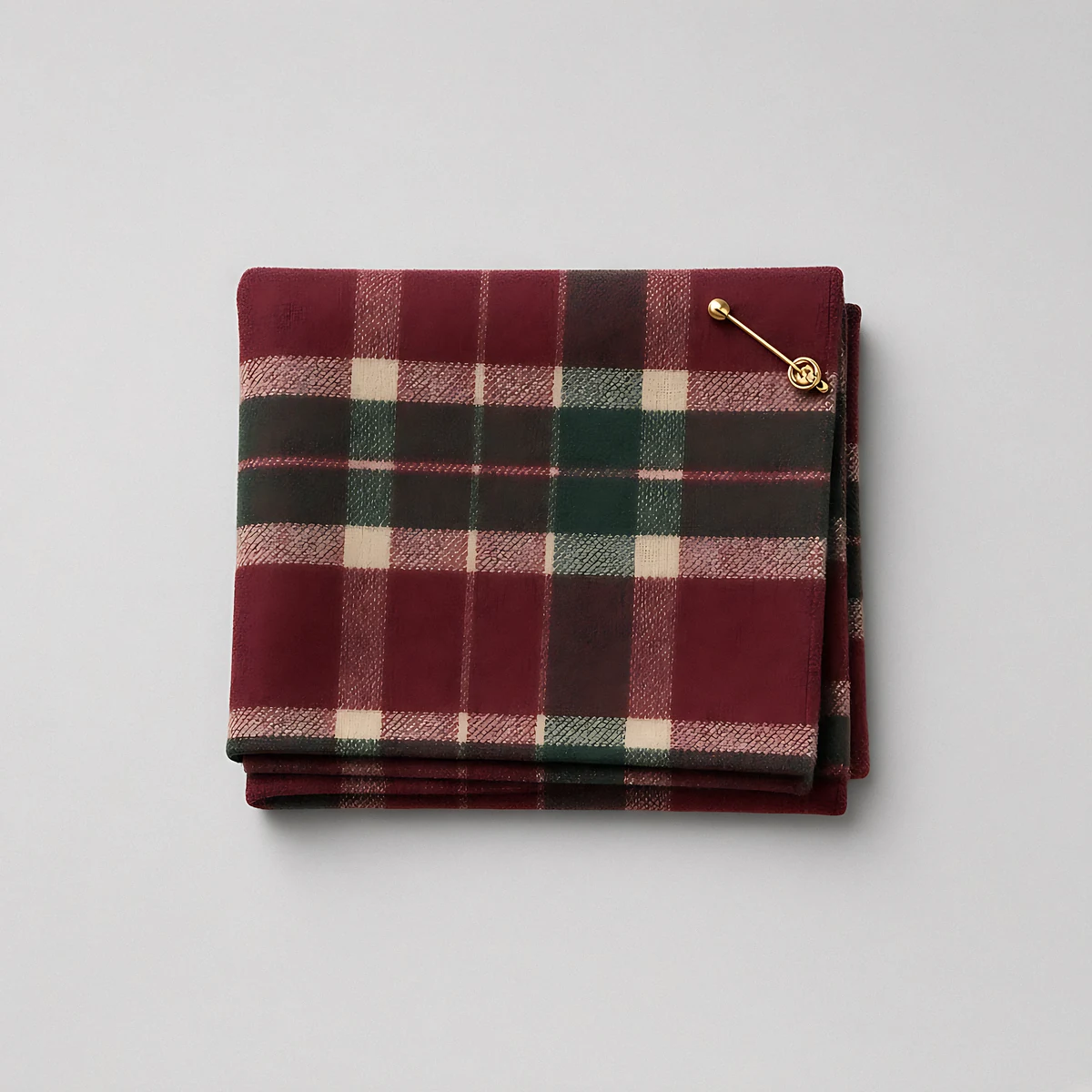 A folded burgundy, cream and green tartan wool scarf with a gold pin on a pale grey background