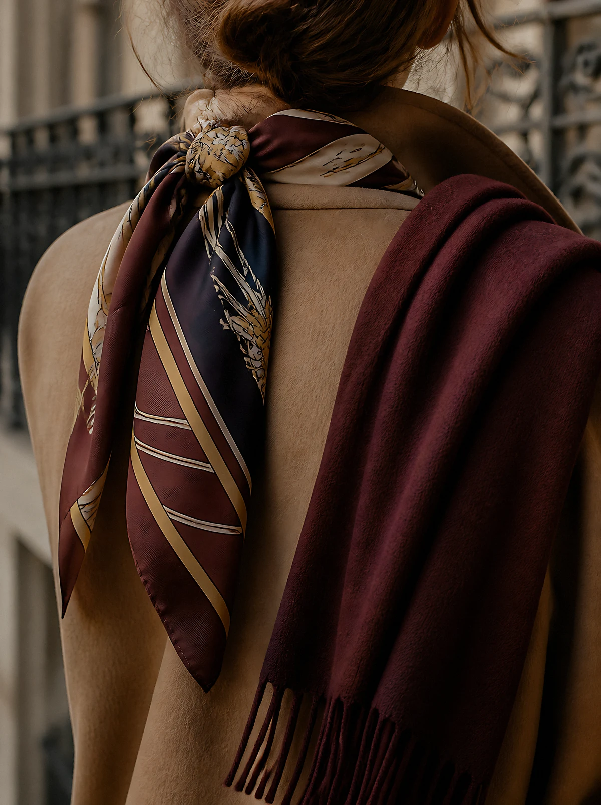 A woman seen from behind in a camel coat, a navy and burgundy striped silk scarf knotted at her neck and a fringed burgundy wool scarf over her shoulder